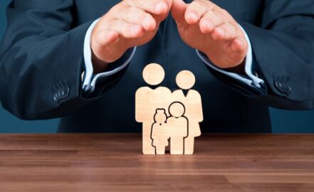 A businessman with a protective gesture over wooden figures representing an insured family