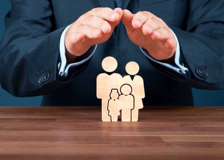A businessman with a protective gesture over wooden figures representing an insured family
