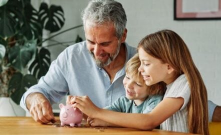 A grandparent and his grandchildren putting money in a piggy bank
