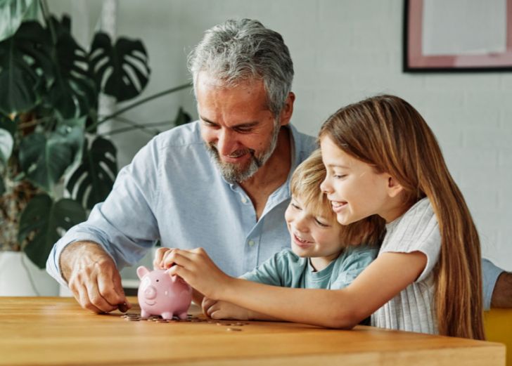 A grandparent and his grandchildren putting money in a piggy bank
